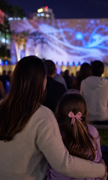 Mother and young daughter watching a nighttime outdoor projection event at SoundScape Park