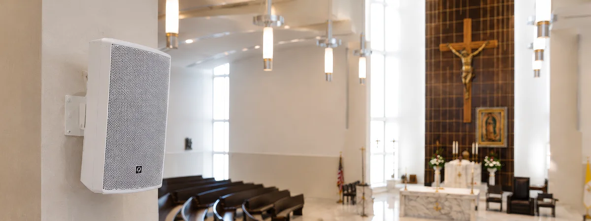 Church Column Speaker Installation White column-mounted church speaker inside a modern sanctuary with curved pews, pendant lighting, altar, crucifix, and worship audio system integration